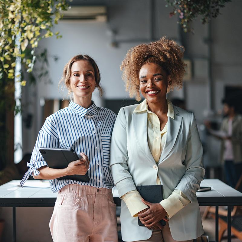 Two Happy Beautiful Businesswomen Looking At Camera While Standing In The Office